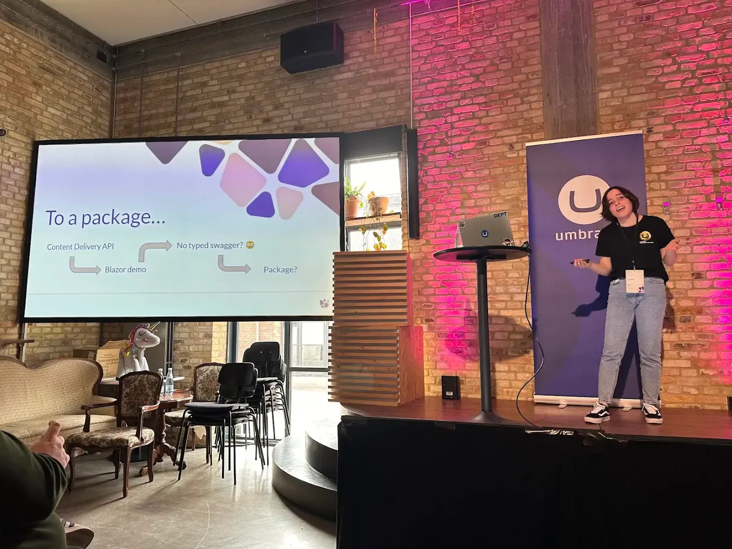 Laura Neto on stage giving her talk. Behind her is a pull-up blue Umbraco banner and a brick wall illuminated by pink lights. To the left is a projector screen showing her slides.